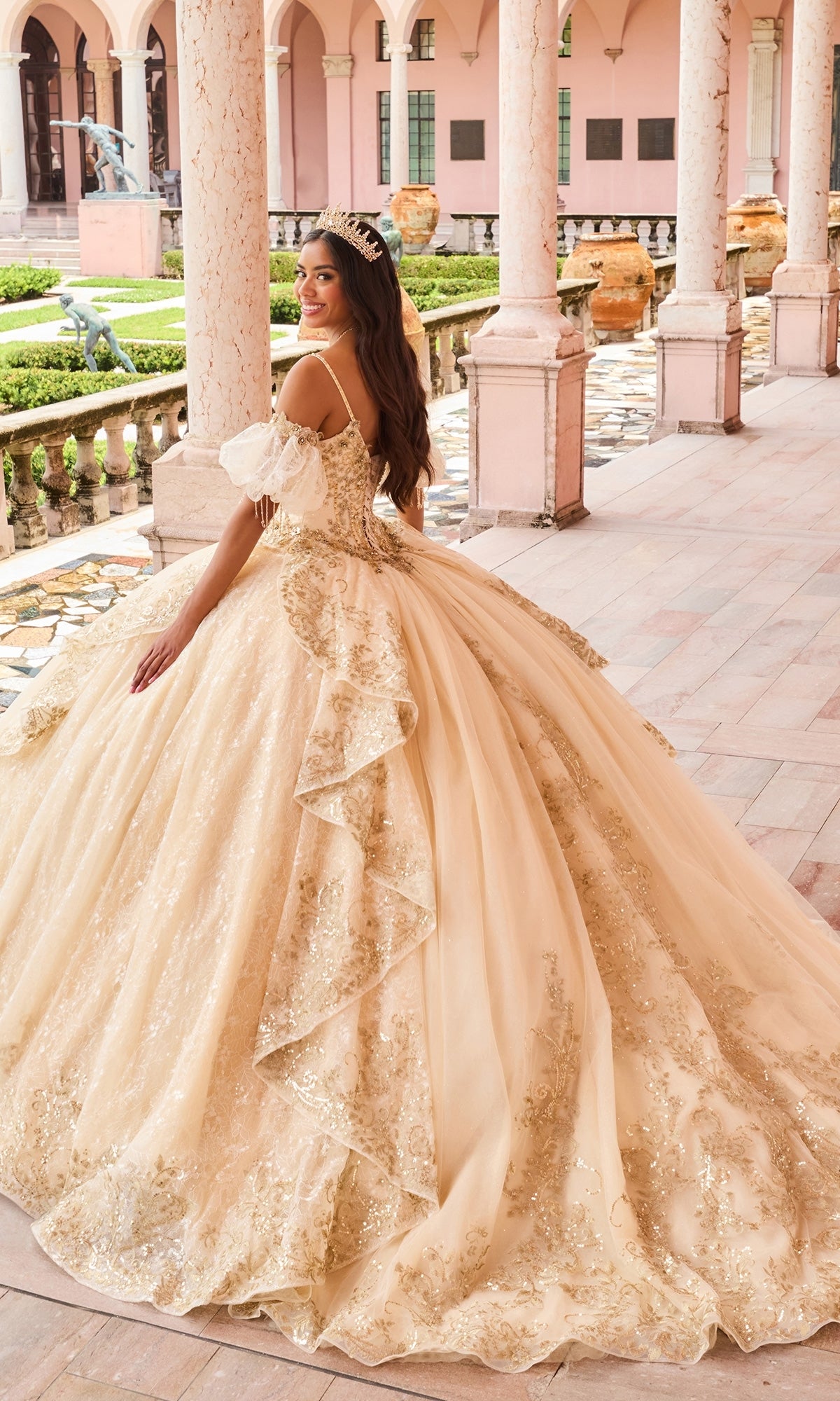 Wearing the Lace Quinceanera Dress: Princesa PR30161, a woman in a gold embellished ball gown with a glitter tulle detachable train and tiara stands in a columned walkway, looking over her shoulder with a smile.